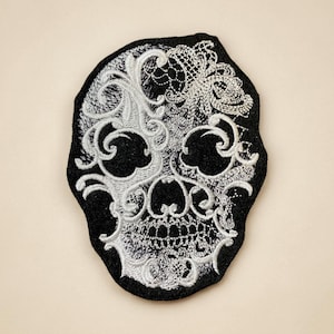 May include: Black skull patch with intricate white lace-like embroidery. The patch features a detailed design of a skull, with the white embroidery creating a striking contrast against the black background. This patch is suitable for clothing or accessories.