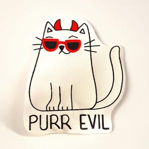May include: White cat-shaped patch with black outline, red devil horns, and red sunglasses. The text "PURR EVIL" is printed in black at the bottom. The patch is on a white background.