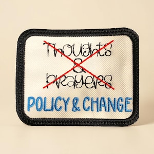 以下が含まれることがあります： クリーム色の背景と黒い縁取りの刺繍パッチ。「Thoughts & Prayers」の文字が赤で消され、その下に青色で「POLICY & CHANGE」と書かれています。