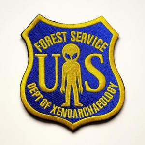 May include: Embroidered patch in a shield shape with a blue background and yellow trim. The patch features the text "FOREST SERVICE" and "DEPT OF XENOARCHAEOLOGY". A yellow alien figure stands between the letters "U" and "S".