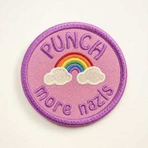 May include: A round, embroidered patch with a pink background and a purple border. The patch features a rainbow, two white clouds, and the text "PUNCH more nazis" in purple.