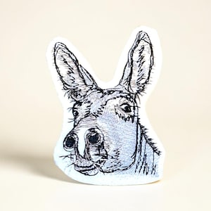 May include: A whimsical donkey-shaped applique with a hand-drawn appearance. The donkey's head is rendered in black and gray, featuring detailed linework and a soft, white background. The applique is likely for crafting or decorative purposes.