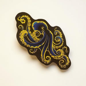May include: Embroidered patch featuring a stylized octopus design. The octopus has blue tentacles and body, with gold detailing. The patch is on a black background and has an irregular shape.