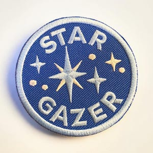 Pode incluir: Patch bordado com fundo azul e as palavras "STAR GAZER" em azul claro. O patch apresenta um design central de estrela e várias estrelas menores em amarelo claro e branco.
