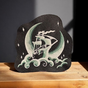 May include: Black embroidered patch featuring a sailing ship set against a crescent moon. The design includes white and green thread, with stars and stylized waves. The patch has a unique, irregular shape.