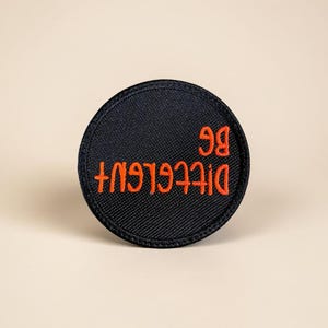 May include: A round, black patch with orange embroidered text that reads "Be Different". The patch has a textured appearance and is likely made of fabric. The text is in a handwritten style.