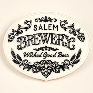 May include: An oval, white patch with black text and decorative elements. The text reads "Salem Brewery" and "Wicked Good Beer." The design includes hops, wheat, and ornate flourishes, creating a vintage aesthetic.