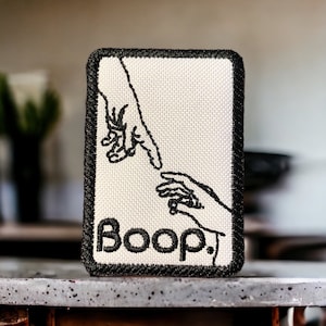 Pode incluir: Um remendo branco bordado com duas mãos que se alcançam. O remendo tem uma borda preta e a palavra "Boop" bordada em preto abaixo das mãos.