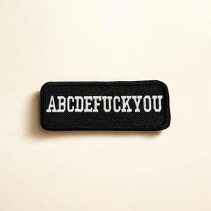 Pode incluir: Um emblema retangular preto bordado com cantos arredondados, apresentando o texto branco ABCDEFUCKYOU numa fonte de estilo bloco em negrito. O emblema tem uma borda preta espessa e uma superfície de tecido texturizado para vestuário.