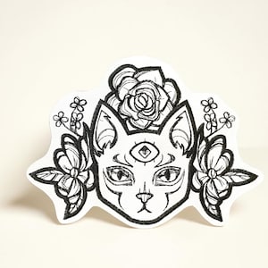 May include: Embroidered patch with a black and white cat design, adorned with flowers and a third eye. The patch features a rose above the cat's head and floral elements on the sides, offering a mystical and decorative appearance.