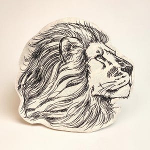 Pode incluir: Uma almofada em forma de leão, de cor creme, com um design bordado a preto detalhado. O perfil do leão é retratado com uma juba fluida e traços faciais. O objeto é decorativo.