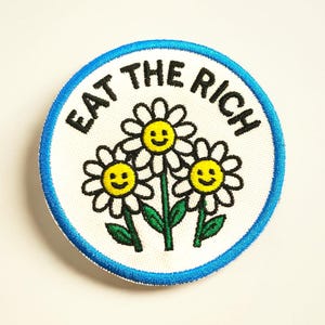 May include: Round patch with a white background, blue border, and the text "EAT THE RICH" in black. The patch features three smiling daisy flowers with yellow centers, white petals, and green stems and leaves.