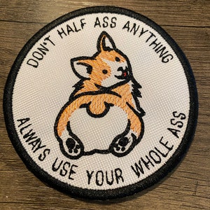 May include: Circular embroidered patch with a corgi dog design, showing its rear end. The patch has the text "DON'T HALF ASS ANYTHING ALWAYS USE YOUR WHOLE ASS" in black, around the edge. The corgi has orange and white fur.