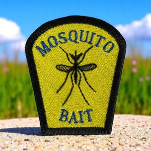 May include: A yellow patch with the words "MOSQUITO BAIT" in blue, featuring a black mosquito illustration. The patch has a black border and is set against a blurred background of green grass and a blue sky.