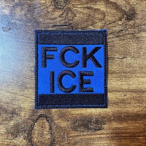 Op de afbeelding: Een vierkante patch met de tekst "FCK ICE" in zwarte blokletters op een blauwe achtergrond. De patch is omrand met zwarte stof en is waarschijnlijk gemaakt van een duurzaam materiaal.