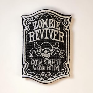May include: Black and white embroidered patch with the words "ZOMBIE REVIVER" in a decorative font. The patch features a skull and crossbones design with the text "EXTRA STRENGTH VOODOO POTION". The patch has a decorative border.