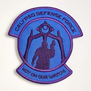 May include: A blue and purple embroidered patch with the text "CALYPSO DEFENSE FORCE" and "NOT ON OUR WATCH!". The patch features a purple silhouette of a figure holding a weapon, with a stylized design above.