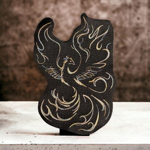 Peut inclure: Un patch en tissu noir avec un motif de phénix brodé en or. Le phénix est représenté avec ses ailes déployées et un motif de flammes tourbillonnantes autour de lui.