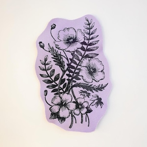 Op de afbeelding: Een lavendelkleurige, onregelmatig gevormde onderzetter met een zwart bloemenontwerp. Het ontwerp bevat gedetailleerde illustraties van bloemen, bladeren en stelen. De onderzetter is waarschijnlijk gemaakt van stof.
