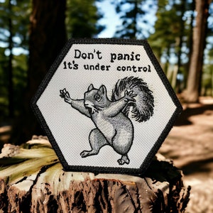 Könnte beinhalten: Ein weißer sechseckiger Patch mit schwarzen Nähten und einem schwarzen Rand. Der Patch zeigt ein Cartoon-Eichhörnchen mit buschigem Schwanz und dem Text "Don't panic it's under control".