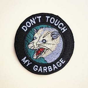 May include: Embroidered patch with a black border and the text "DON'T TOUCH MY GARBAGE" in white. The patch features a cartoon opossum with a gray and white face, open mouth, and red tongue, set against a teal and black background.