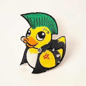 May include: Embroidered patch featuring a yellow duck with a green mohawk, a safety pin, and a cigarette. The duck is wearing a black jacket with a star and peace sign. The patch has a white background.