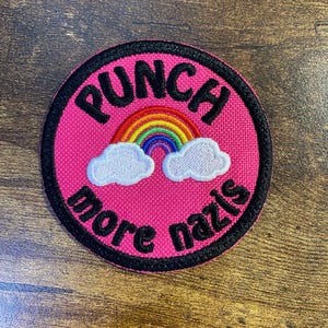 May include: A round, pink patch with black trim. The patch features the text "PUNCH more nazis" in black, arched around a rainbow and two white clouds.