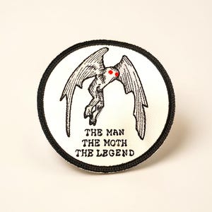 May include: A round patch featuring a black and white illustration of a moth-like creature with red eyes. The text "THE MAN THE MOTH THE LEGEND" is printed below the image. The patch has a black border.