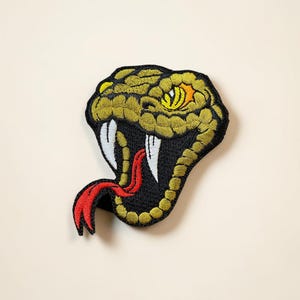 May include: Embroidered patch featuring a snake head with an open mouth. The snake's head is primarily olive green and black, with a yellow eye and white fangs. A red tongue extends from the mouth. The patch is likely for clothing or accessories.