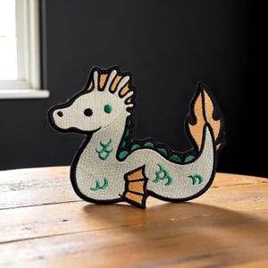 以下が含まれることがあります： ライトグリーン、オレンジ、黒色の海のヘビの刺繍パッチ。 海の生き物は、長い湾曲した体とヒレのような尾、詳細な頭を持っています。 衣類やアクセサリー用です。