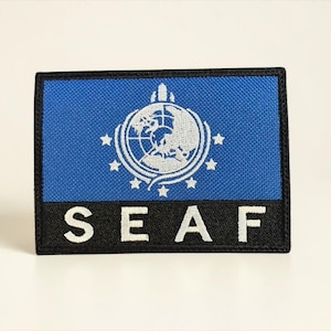 May include: A rectangular patch with a blue background and a white emblem featuring a globe and stars. The patch has a black border and the letters "SEAF" in white.