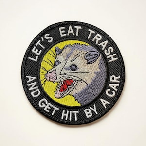 May include: Embroidered patch with a circular design featuring a possum with an open mouth. The text "LET'S EAT TRASH AND GET HIT BY A CAR" is embroidered around the possum in white letters on a black background.