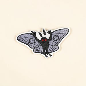 Op de afbeelding: Geborduurde patch met een gestileerde mot met een zwart lichaam, rode ogen en grijze vleugels. De mot heeft zijn armen omhoog en is wit omlijnd. De vleugels hebben een gedetailleerd patroon. Voor kleding of accessoires.