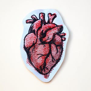 May include: Embroidered patch featuring a detailed anatomical heart design. The heart is rendered in shades of red and black, with intricate line work. The patch has a light blue border and is likely intended for application on clothing or accessories.