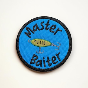 Könnte beinhalten: Gestickter Aufnäher mit blauem Hintergrund und schwarzem Rand. Der Aufnäher zeigt die Worte "Master Baiter" in schwarzer Schrift, mit einem gelb-grünen Angelköder in der Mitte.