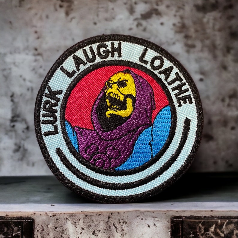 May include: A black and white embroidered patch with a circle design. The circle features a red background with a purple and blue figure with a yellow skull and the text "Lurk Laugh Loathe" around the circle.