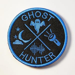 Puede incluir: Parche redondo negro con texto y gráficos bordados en azul. El texto "GHOST HUNTER" está bordado en el borde. El diseño incluye un fantasma, luna, estrellas, linterna y ondas de sonido.