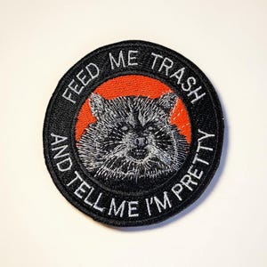 Könnte beinhalten: Runder, schwarzer Aufnäher mit einem Waschbären-Design. Der Aufnäher hat den weißen Text "FEED ME TRASH AND TELL ME I'M PRETTY" um das Waschbären-Bild, das sich vor einem roten Hintergrund befindet.