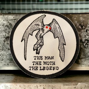 Könnte beinhalten: Ein weißer gestickter Aufnäher mit schwarzem Rand. Der Aufnäher zeigt eine schwarz-weiße Illustration einer mottenartigen Kreatur mit roten Augen. Der Text "THE MAN THE MOTH THE LEGEND" ist unterhalb der Kreatur aufgedruckt.