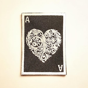 May include: Black rectangular patch with a white embroidered heart design. The patch features the letter "A" in the top left corner and the bottom right corner. The heart is filled with intricate swirls and floral patterns.