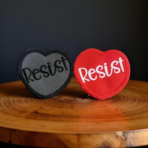 May include: Two heart-shaped patches. One is grey with the word "Resist" in white script. The other is red with the word "Resist" in white script. Both patches are on a wooden surface.