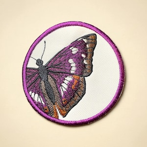 Puede incluir: Parche bordado con una mariposa morada con detalles marrones y blancos. La mariposa está sobre un fondo blanco, enmarcada por un borde circular morado. Diseñado para aplicar en ropa o accesorios.