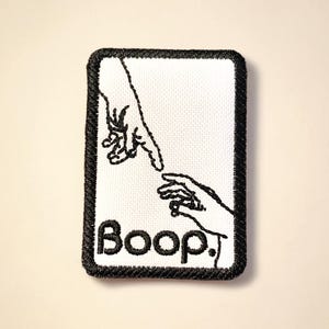 May include: Embroidered patch with a black border and a white center. The design features two hands nearly touching, with the word "Boop!" in black script below. A fun, quirky accessory.