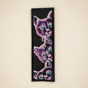 May include: Embroidered patch featuring three stylized cat faces in shades of purple, with green eyes, on a black background. The patch has a rectangular shape with a black border. The cats are stacked vertically.