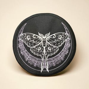 May include: A round, black patch featuring a detailed white moth design with a crescent moon and intricate lace-like patterns. The moth has detailed wings and body. The patch is likely for clothing or accessories.