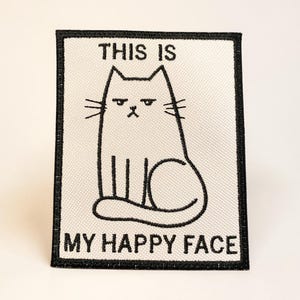 May include: A rectangular patch with a black border and a white background. The patch features a black illustration of a cat with a grumpy expression. The text "THIS IS" is above the cat, and "MY HAPPY FACE" is below.