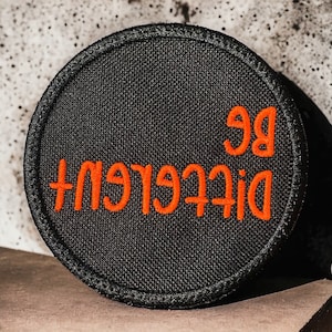 May include: Black round patch with orange embroidered text that reads "Be Different".