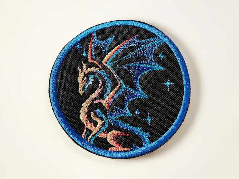 May include: Embroidered patch featuring a detailed dragon design in shades of blue, red, and gold against a black background. The patch has a blue border and includes small star accents, suitable for jackets, bags, or other fabric items.