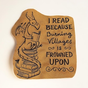Könnte beinhalten: Senffarbener Aufnäher mit einem Drachen, der ein Buch auf einem Bücherstapel liest. Der Drache trägt eine Brille und hat Flügel. Der Text lautet: "I READ BECAUSE Burning Villages is FROWNED UPON."
