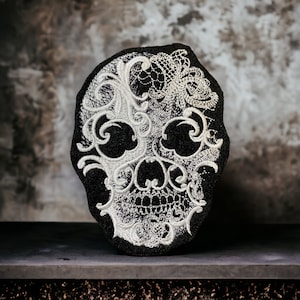 May include: A black and white embroidered skull patch with a floral design. The skull is outlined in white thread and the floral design is intricate and detailed.